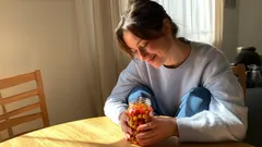 Dívka u stolu drží sklenici plnou barevných bonbonů v slunečně osvětlené místnosti.